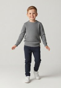 Szary dziany sweter z okrągłym dekoltem, zestawiony z ciemnoniebieskimi spodniami. Dziecko nosi białe trampki, uśmiecha się, na neutralnym tle.