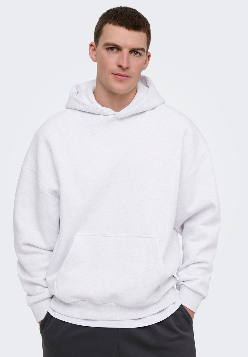 Jeune homme portant un sweat à capuche blanc uni et un pantalon noir, debout les mains dans les poches devant un fond gris clair.