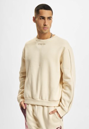 Beige Sweatshirt mit langen Ärmeln, gerippten Bündchen und rundem Ausschnitt. Mit "Thug Life"-Stickerei auf der Brust und Reißverschlussdetail an der Seite.