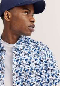 Kurzarmhemd mit Knöpfen, das ein blaues Blumenmuster auf weißem Hintergrund zeigt, mit braunen Knöpfen und einem klaren Kragen.