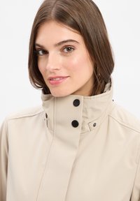 Beige Regenjacke mit hohem Kragen, schwarzen Druckknöpfen und einer glatten, wasserabweisenden Textur.
