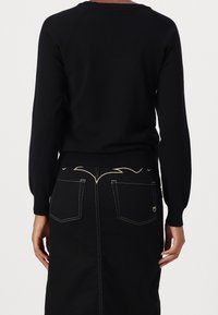 Personne portant un pull noir à manches longues et une jupe noire avec des coutures blanches contrastantes et un détail décoratif sur la poche arrière, vue de dos.