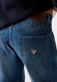 Dunkelblaue Denim-Jeans mit einem Leder-Guess-Logo-Patch auf der Rückseite. Verfügt über zwei Gesäßtaschen mit sichtbaren Nähten und Logo.