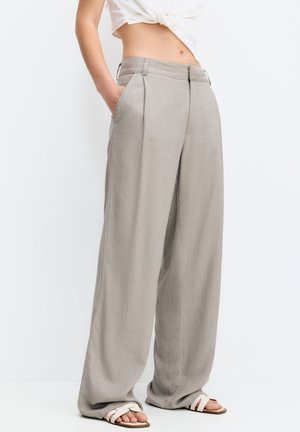 Vrouw draagt een beige broek met hoge taille en wijde pijpen met zakken, gecombineerd met witte sandalen met bandjes en een wit cropped topje met strik.