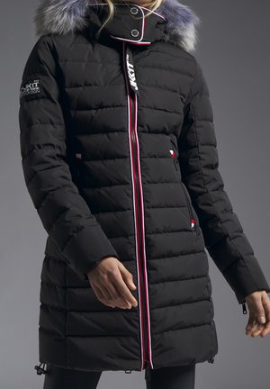 Czarna pikowana zimowa kurtka z kapturem wykończonym futerkiem, czerwono-biały suwak w paski oraz naszywka z logo na rękawie.