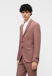 Νεαρός άνδρας με σγουρά μαλλιά που φοράει ένα κοστούμι δύο κουμπιών σε ρόζ ξεθωριασμένο χρώμα και λευκό πουκάμισο, στέκεται μπροστά σε απλό ανοιχτό φόντο.