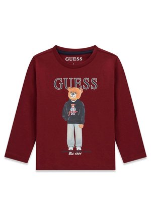Chemise à manches longues marron avec un ours portant un sweat à capuche noir et un pantalon gris, texte "GUESS" au-dessus, "Est. 1981" en dessous.