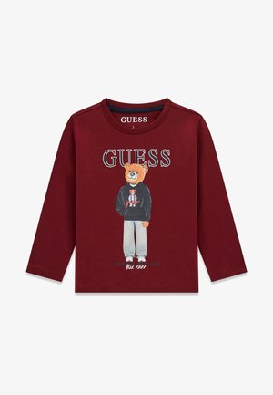 Koszula z długim rękawem w kolorze bordowym z niedźwiedziem w czarnej bluzie z kapturem i szarych spodniach, napis "GUESS" u góry, "Est. 1981" na dole.
