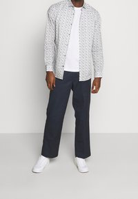 Mężczyzna w białej koszuli z wzorem na guziki, białym podkoszulku, granatowych spodniach i białych trampkach stojący na jednolitym tle.