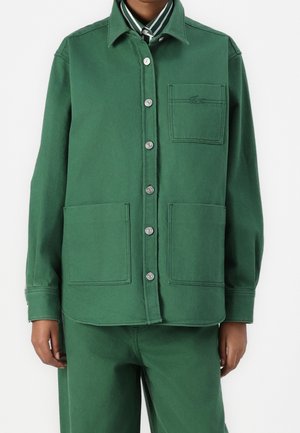 Personne portant une veste verte à boutons avec trois grandes poches devant et un pantalon assorti, debout sur fond blanc.