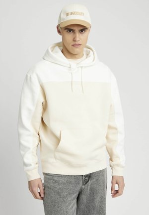 Hoodie crème avec une poche avant et un cordon de serrage, présente une section supérieure plus claire. Porté avec des jeans gris déchirés et une casquette beige avec l'inscription "SOIS LÉGENDAIRE."