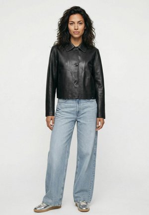 Frau trägt schwarze Lederjacke, hellblaue Jeans mit weitem Bein und silberne Sneakers und steht vor einem einfarbigen weißen Hintergrund.