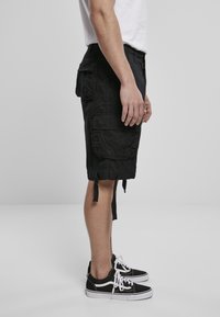 Man som bär svarta cargoshorts, vit T-shirt och svarta låga sneakers med vita snören, stående i profil mot en enkel bakgrund.