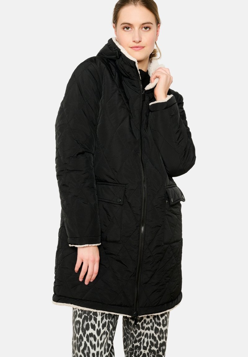 Čierny prešívaný dlhý kabát s kapucňou, ktorý má jemnú smotanovú podšívku, dve predné vrecká a zipsové zapínanie. Rukávy sú manžetové s smotanovým lemom.