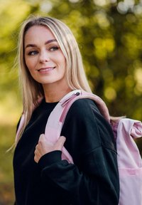 Růžový batoh s hladkým povrchem a polstrovanými popruhy, nošený přes černou mikinu. Měkké rozostřené zelené pozadí.