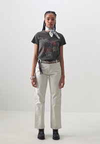 Grå grafisk t-shirt med en design av en svart panter, beige rak benbyxor, svarta stövlar och en vit scarf knuten runt halsen.