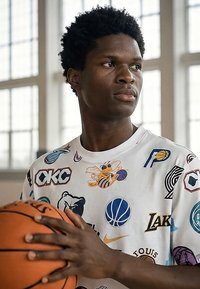 Mladý muž držiaci basketbal, oblečený v bielom tričku s rôznymi logami tímov NBA, stojaci vo vnútri pri veľkých oknách, pozerá sa do strany.