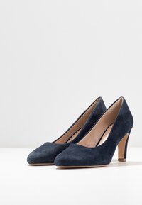 Laivastonsiniset mokkanahkasaappaat, joissa on pyöreä varvas ja keskipituinen laatikkokorko, erottuvat sileällä pinnalla ja minimalistisella muotoilulla.