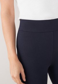 Tamno plave, leggins hlače visokog struka s glatkim, rastezljivim materijalom i teksturiranim završetkom. Prikaz bliže pojasa i ruke.