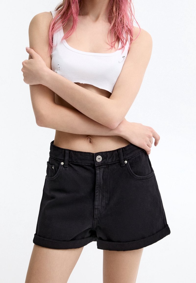 Personne aux cheveux roses porte un débardeur blanc court et un short en denim noir taille haute avec les ourlets retroussés, les bras croisés sur la poitrine.