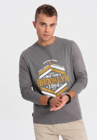 Graues Langarm-T-Shirt mit einem grafischen Druck in Gelb und Weiß, der den Text "BROOKLYN 1964" zeigt. Es hat einen Rundhalsausschnitt und eine weiche Textur.