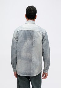 Džínsová košeľa vo svetlo sivej farbe s textúrovaným vzorom, predné zapínanie na gombíky a dlhé rukávy. Viditeľné švy a dizajn zadného sedla.