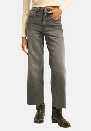 Femme portant un jean gris taille haute à jambes larges, des bottines noires et un top court beige à manches longues, une main dans la poche.