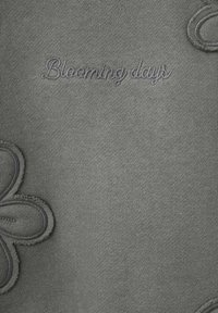 Tessuto grigio con una texture liscia e motivi floreali ricamati. La frase "Blooming days" è cucita con un filo dello stesso colore.