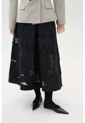 Schwarze Midi-Rock mit strukturiertem Blumenmuster, über einer durchsichtigen Basis geschichtet. Das Outfit umfasst ein graues Blazer und schwarze spitze Ballerinas.
