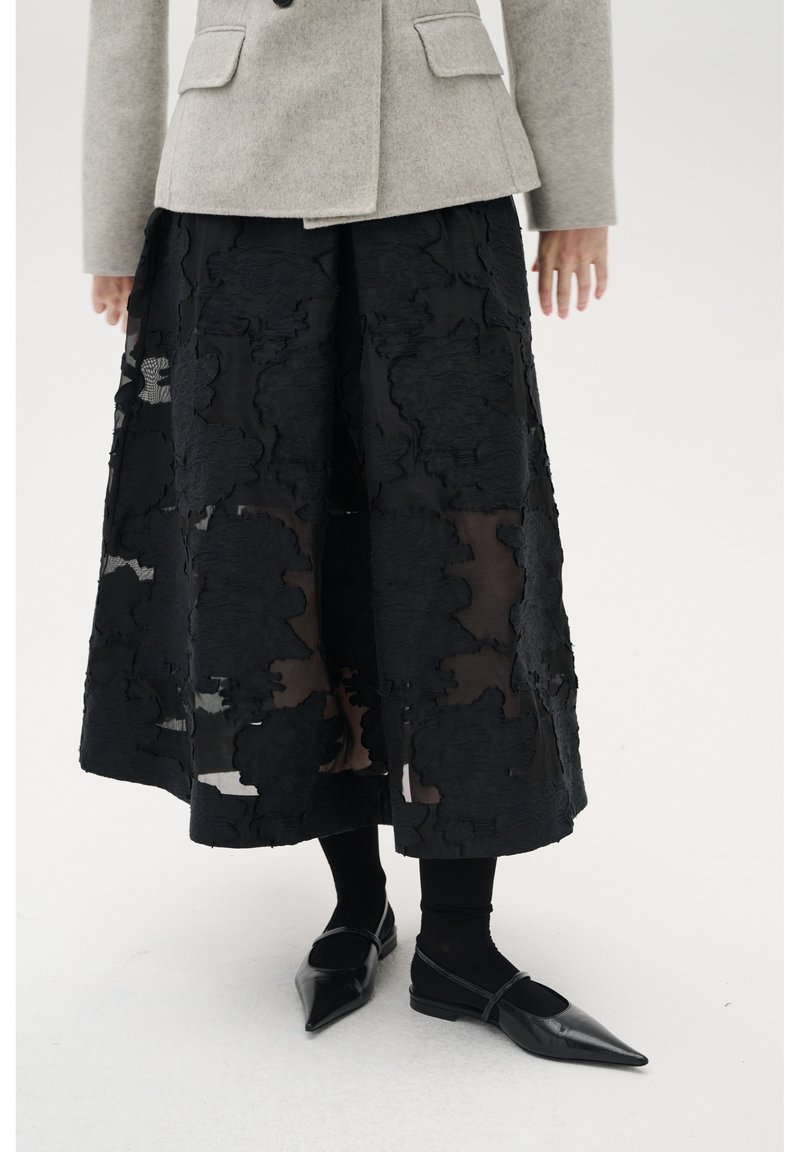 Sort midi-nederdel med struktureret blomster mønster, lagdelt over en gennemsigtig bund. Outfittet inkluderer en grå blazer og sorte spidse flade sko.
