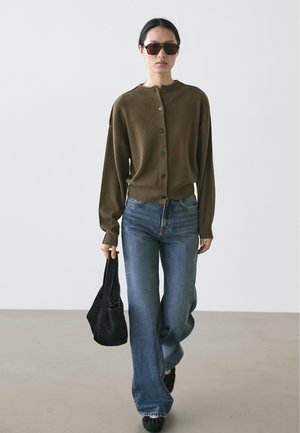 Bruine gebreide cardigan met knopen, gecombineerd met lichtblauwe wijde jeans. Accessoires zijn onder andere een zwarte zonnebril en een kleine zwarte tas.