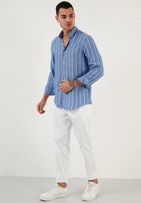 Camicia button-up azzurro chiaro con righe verticali bianche, maniche lunghe e colletto scuro a contrasto, abbinata a pantaloni bianchi e sneakers.
