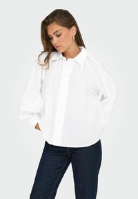 Bílá bavlněná oversized košile s špičatým límcem, zapínáním na knoflíky a nabíranými rukávy. Má volný střih a jemný lesk. Nosič má na sobě tmavé džíny.