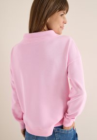 Lichtroze sweatshirt met hoge kraag, lange mouwen en ribgebreide manchetten, gedragen met blauwe denim jeans. Soepele textuur en losse pasvorm.