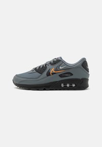 Grå Nike Air Max-sneaker med en texturerad mesh-övre del, svarta accenter och en orange swoosh, med en synlig luftkuddenhet i sulan.