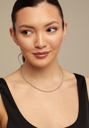 Mujer de cabello oscuro con una blusa sin mangas negra que lleva un collar plateado con cuentas, mirando hacia su izquierda con una leve sonrisa sobre un fondo neutro.