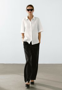Camicia bianca a maniche corte in lino, pantaloni neri larghi e sandali neri chunky. Gli accessori includono occhiali da sole e orecchini a cerchio.