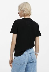 Black cotton t-shirt with short sleeves, featuring a subtle graphic detail on the back. Paired with light blue denim shorts.