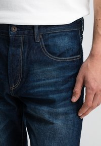 Donkerblauwe spijkerbroek met een rechte pasvorm, subtiele vervaging, contrasterende stiksels en een metalen knoop bij de tailleband.