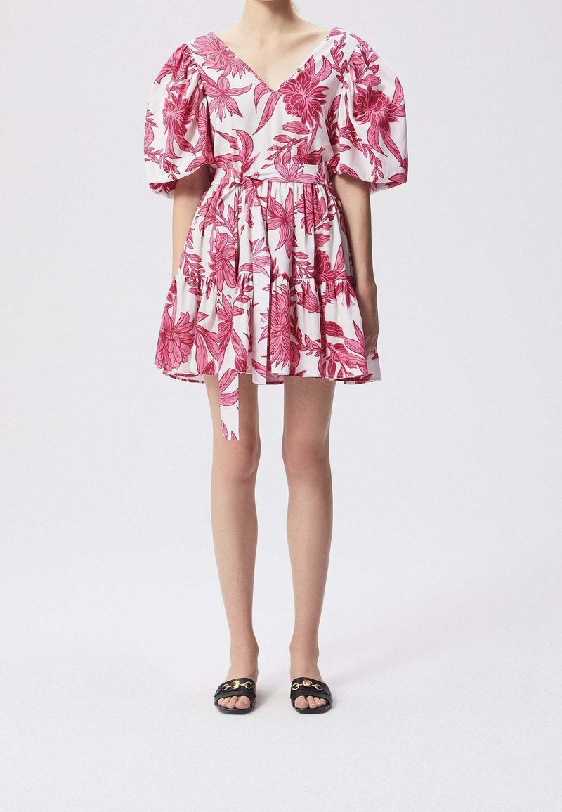 Woman wearing a short white dress with pink floral print, puffed sleeves, a waist tie, and black open-toe slip-on sandals with gold hardware.