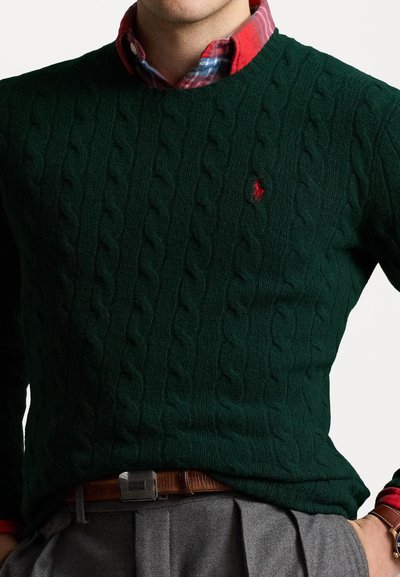 Πράσινο πουλόβερ με πλέξιμο και λαιμόκοψη, που διαθέτει ένα μικρό κόκκινο λογότυπο στο στήθος. Φοριέται πάνω από ένα καρό πουκάμισο σε κόκκινες και μπλε αποχρώσεις.