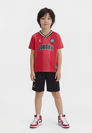 Fiatal fiú piros Jordan kosárlabdamezben, fekete rövidnadrágban és piros-fehér sportcipőben, sima fehér háttér előtt áll.