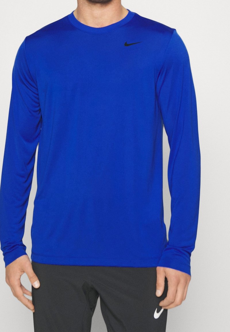 Man med en enkel blå långärmad Nike-tröja och svarta Nike-byxor mot en enfärgad ljus bakgrund.