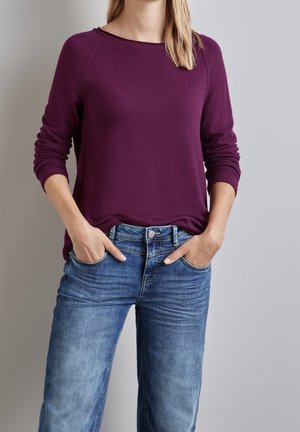 Vrouw met een lange paarse trui met lange mouwen en een blauwe spijkerbroek met handen in de zakken tegen een grijze achtergrond.