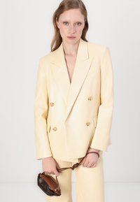 Blazer doppiopetto in ecopelle gialla chiara, dotato di grandi bottoni rotondi dorati e revers a punta. Abbinato a pantaloni coordinati e a una borsa marrone.