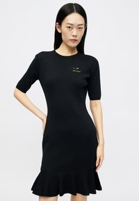 Vestido preto de malha com mangas curtas, corpete justo e barra rodada com folhos. Apresenta um discreto logótipo dourado no peito. Textura suave.