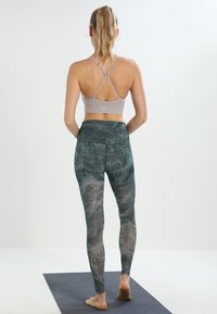 Grijze sportbeha met kruisende rugbanden, gecombineerd met groene legging met een wit abstract patroon. Model staat op een yoga mat.