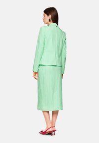 Mint green pleated suit with a fitted jacket and knee-length skirt. Worn with red slingback heels. Back view, smooth texture, simple design.