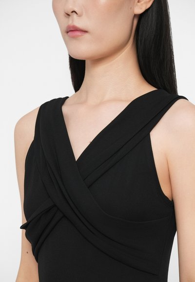 Γυναίκα φορεμένη με μαύρο αμάνικο φόρεμα με φαρδιές, επικαλυπτόμενες τιράντες που σχηματίζουν ντεκολτέ σε σχήμα V, αποκαλύπτοντας το λαιμό και τους ώμους.