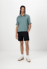 Hellblaues Strick-Polohemd mit kurzen Ärmeln und weißen Akzenten; kombiniert mit schwarzen Shorts und hellen Sneakers, steht vor einem schlichten Hintergrund.